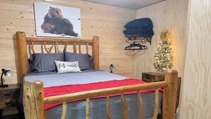 A wooden bed with a red blanket, a pillow with text, a wall-mounted coat rack, a small Christmas tree, and a bear picture on the wall.
