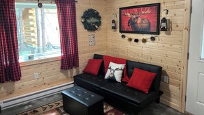 A cozy cabin interior with a black sofa, red cushions, a framed moose picture, and a 'Alaska' sign on the wall.
