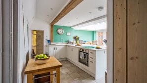 Une cuisine avec des meubles blancs, une table en bois sur laquelle se trouve un saladier rempli de fruits, une horloge au mur et, à travers une ouverture, on aperçoit une autre pièce dont le mur est en briques.