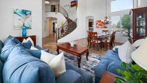 Un salón amplio con un sofá azul, una zona de comedor con sillas de madera, una escalera que sube a un nivel superior y un ventanal que proporciona luz natural.