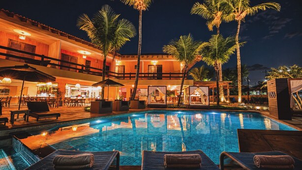 A nighttime pool area with lounge chairs, palm trees, and a bar area. - LUNA Hotel Boutique (Guarujá)