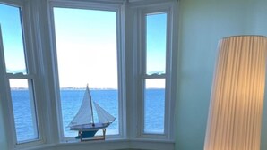Habitación con vista al mar a través de tres ventanas, una silla blanca pequeña, una silla azul, una lámpara y un reposapiés negro con un libro encima.
