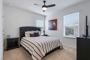A bedroom with a tufted headboard, a striped bedspread, a ceiling fan, a nightstand, a bedside table, a framed artwork, a window with blinds, and a TV stand.