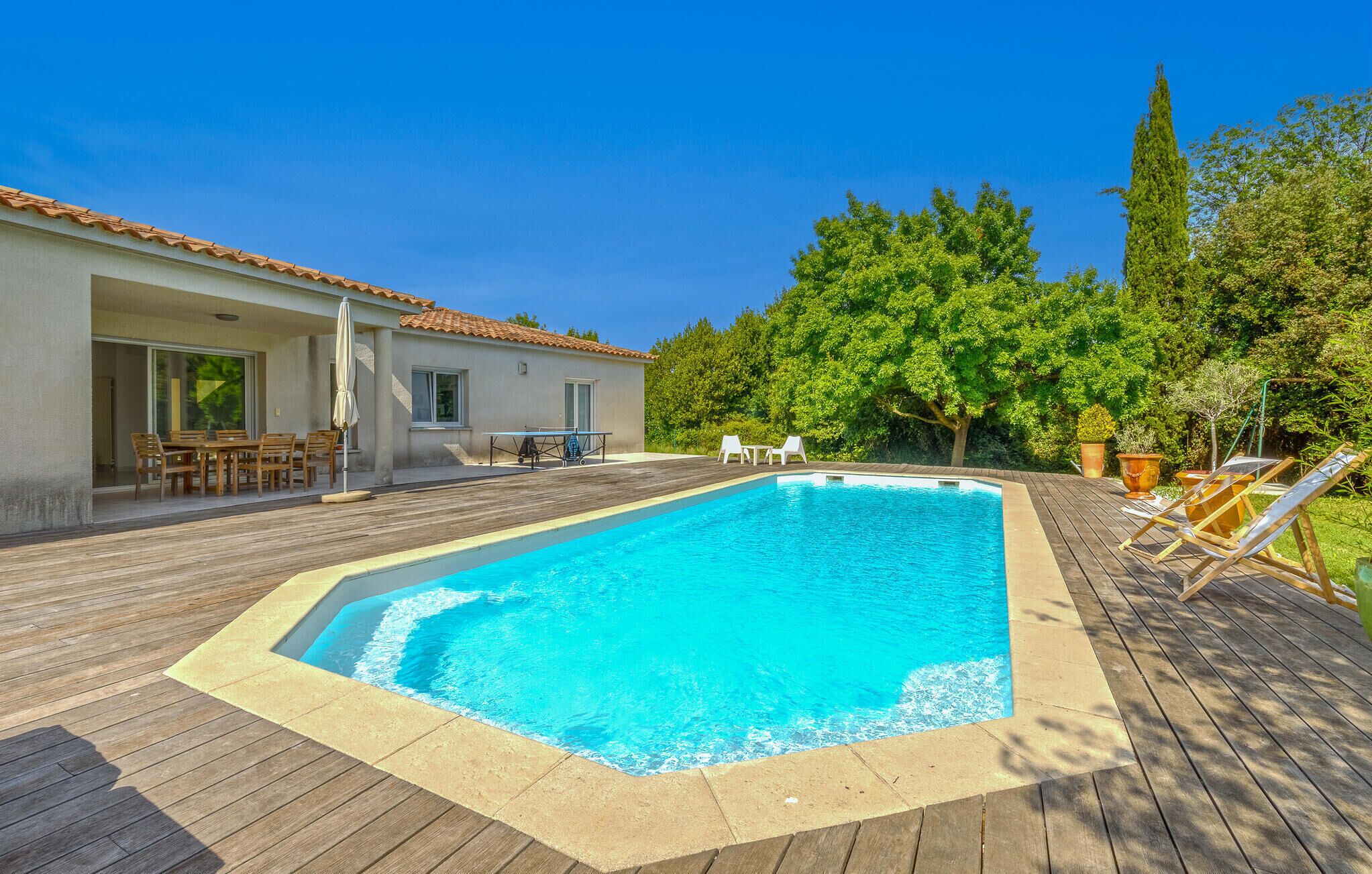 Ein Schwimmbecken mit Holzterrasse, eine überdachte Terrasse mit Gartenmöbeln und ein Haus mit Ziegeldach.