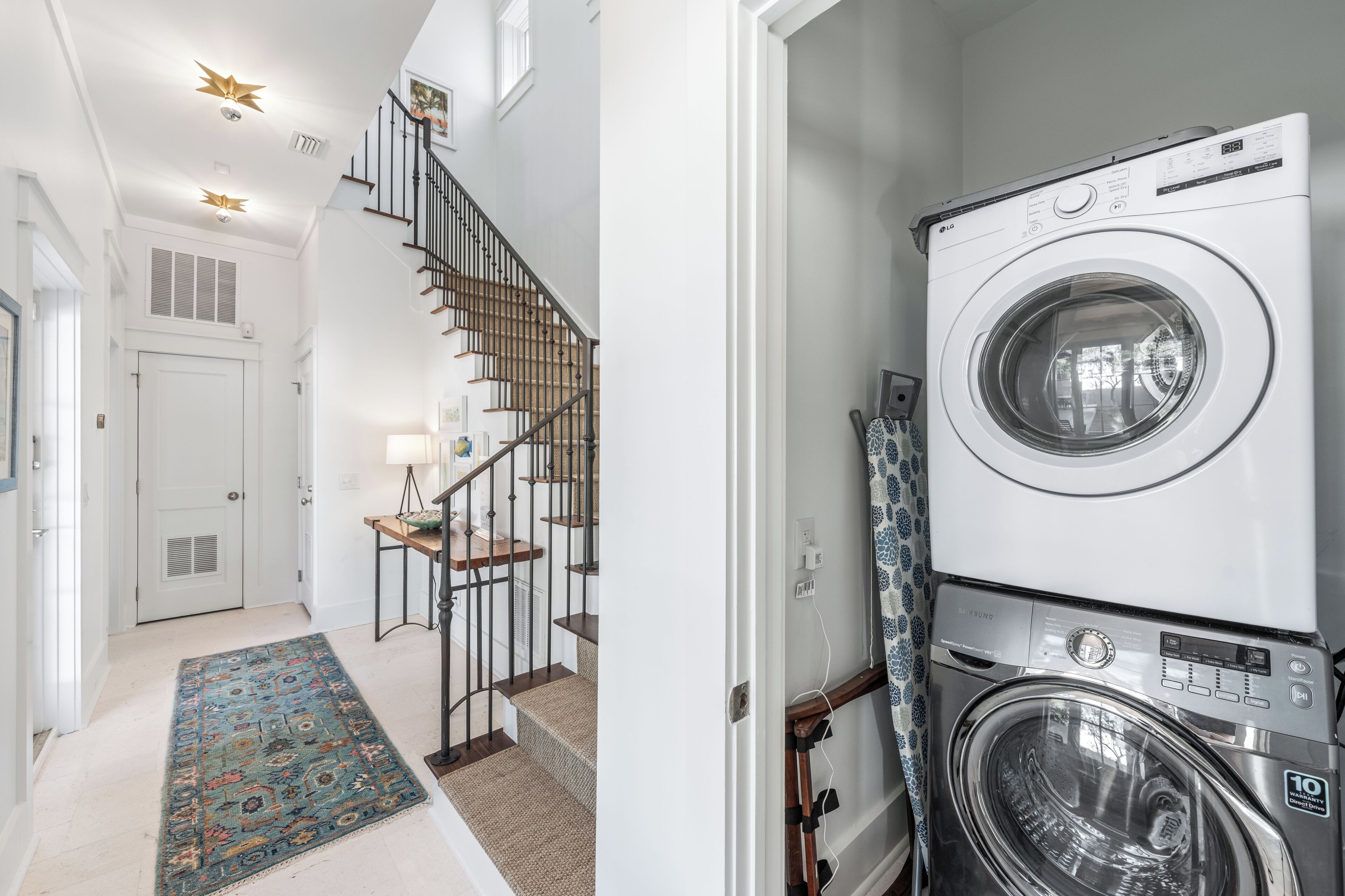 A laundry room with a stacked washer and dryer, a wooden staircase with a railing, and a patterned rug.