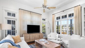 Ein Wohnzimmer mit Flachbildfernseher, weißen Möbeln, Deckenventilator und großen Fenstern.