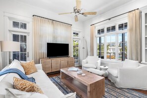 A living room with a flat-screen TV, white furniture, a ceiling fan, and large windows. - Wave Jumper | Cottage in Rosemary Beach with Access to Rosemary Beach, Pools, Tennis, and Gym (Rosemary Beach)
