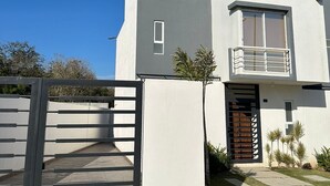 A modern two-story house with a flat roof, a balcony on the second floor, and a gated entrance.