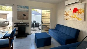 A modern living room with a blue sofa, a dining table with chairs, and a TV stand. There is a painting on the wall and an air conditioning unit above it.