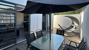An outdoor patio area with a glass table and chairs, a large umbrella, and a view of a modern house.