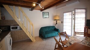 Une cuisine équipée d’un four à micro-ondes, d’un lave-linge et d’un escalier. On y trouve un canapé vert et une table en bois avec des chaises. Une lampe est posée sur une table d’appoint, et un téléviseur est installé sur un meuble.