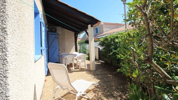 Un espace extérieur abrité, aménagé avec une table et des chaises, une porte bleue et un jardin avec des plantes et un arbre.