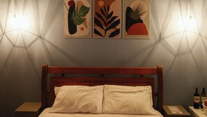 Una camera d'albergo con una testiera in legno, un letto con lenzuola bianche e tre dipinti astratti appesi al muro. Si notano due comodini con lampade e una bottiglia di vino su uno di essi.