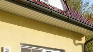 Una casa amarilla con un cartel con el número 26, una ventana, una mesa con sillas y una cesta de fruta.