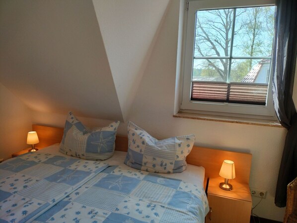 Un dormitorio con cama, dos lámparas de noche, una ventana con persianas y vistas a árboles y un edificio.