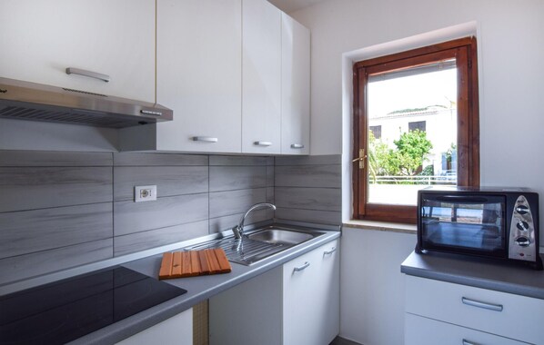 Une cuisine compacte avec des meubles blancs, un four à micro-ondes encastré et une fenêtre donnant sur un immeuble et des arbres.