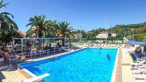 A swimming pool with clear blue water, surrounded by lounge chairs and umbrellas, with palm trees and a hilly landscape in the background.