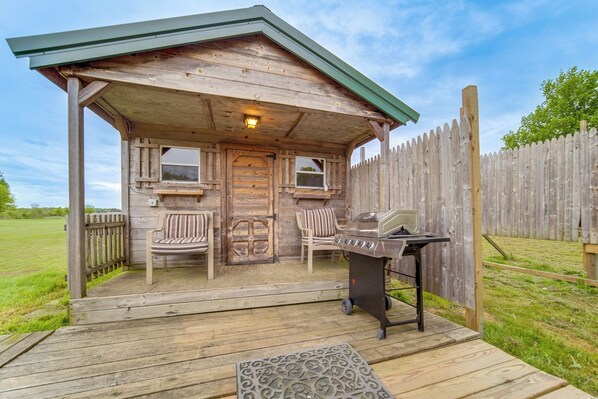 A wooden cabin with a porch, two chairs, a grill, and a small table.