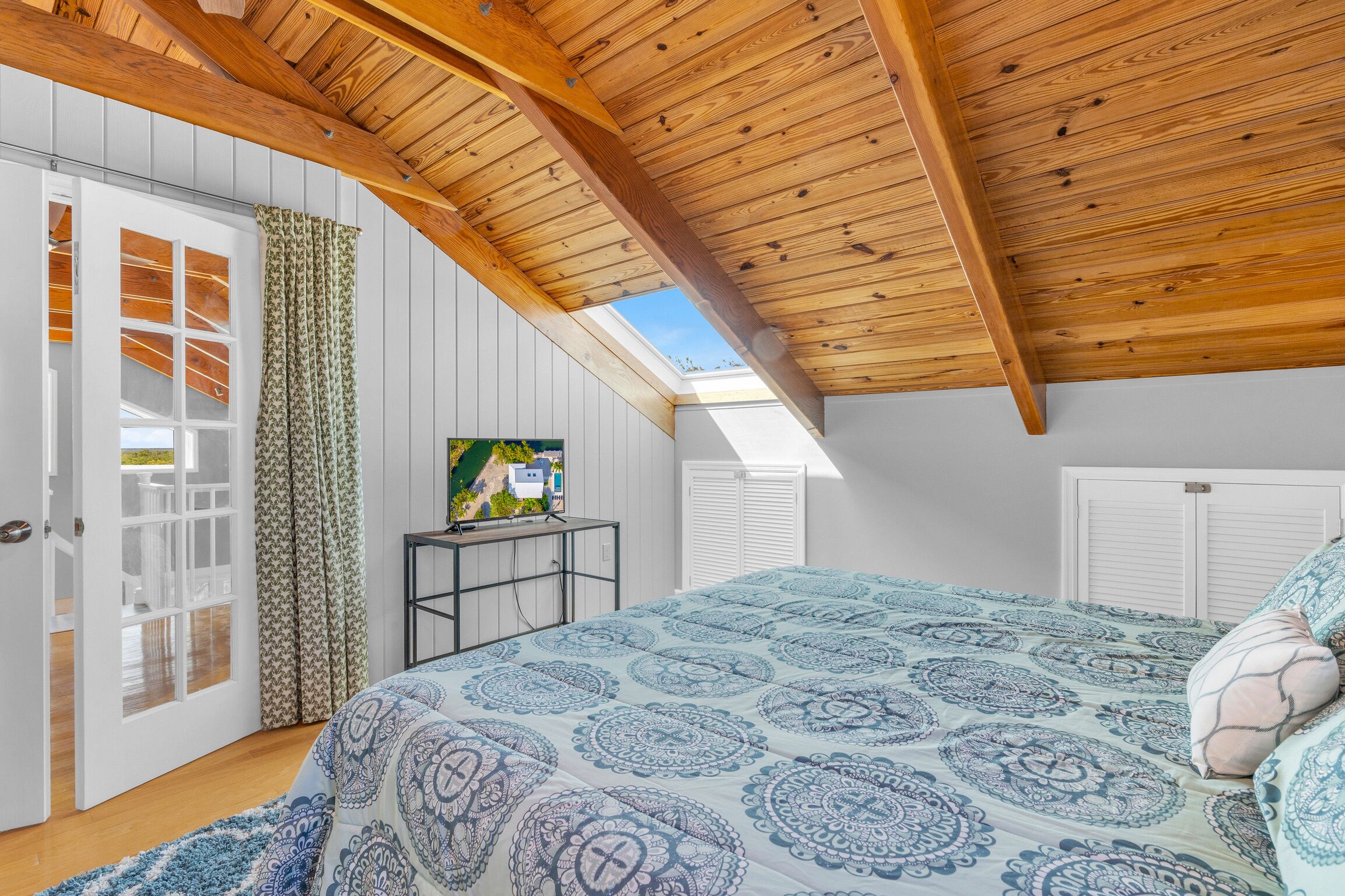 A bedroom with a sloped ceiling, a bed with patterned bedding, a TV on a stand, and a view of the outdoors through a window.