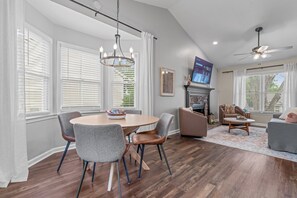 Een ruime woonkamer met een open haard, een eethoek met een ronde tafel en stoelen, en een zithoek met een bank en fauteuils.