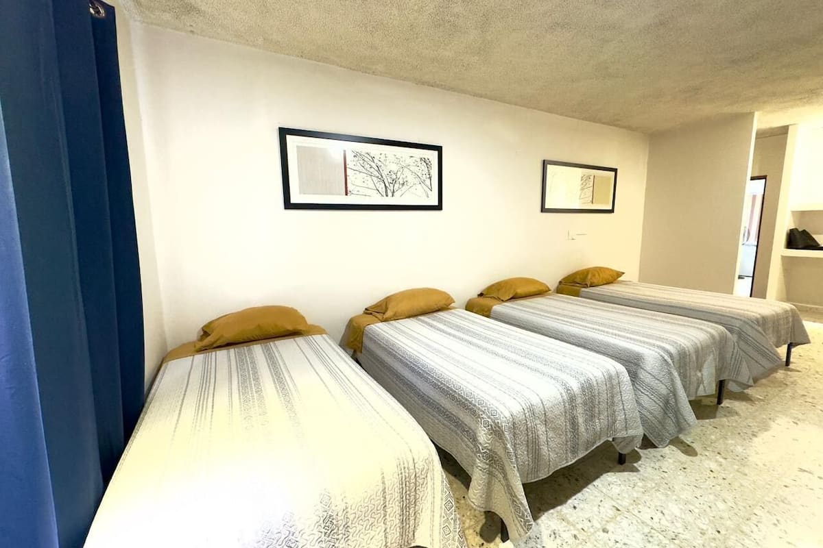 A room with four single beds aligned against a wall, each with a patterned bedspread and a pillow. There are two framed pictures on the wall above the beds.