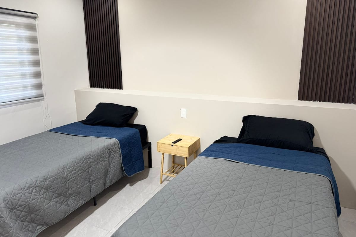 A room with two beds, each with a grey quilted mattress and a blue blanket. There is a small wooden nightstand between the beds with a box on it.
