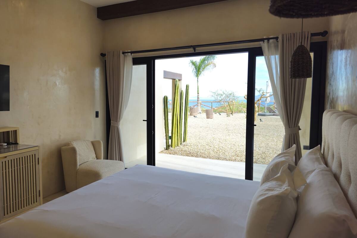 A hotel room with a large bed, a TV mounted on the wall, and a view of a desert landscape with cacti.