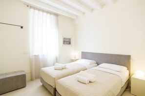 A hotel room with two beds, a bedside table, a lamp, a window with curtains, and a painting on the wall.