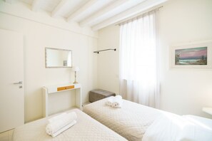 A hotel room with two beds, a bedside table, a mirror, a painting, and a window with curtains.