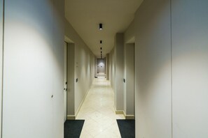 Corredor longo e estreito de um hotel, com portas de ambos os lados, piso ladrilhado e teto com iluminação embutida.