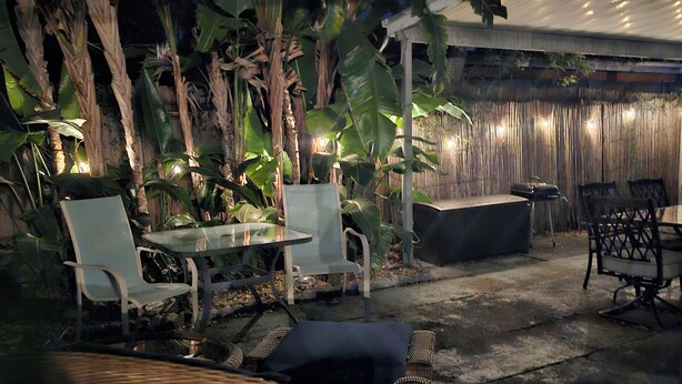 An outdoor patio with wicker furniture, a table, and chairs. There are plants and a bamboo fence in the background.