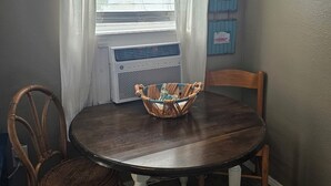 A dining area with a round table, two chairs, a wicker basket, a window with blinds, a wall-mounted sign, and a wall-mounted mirror.