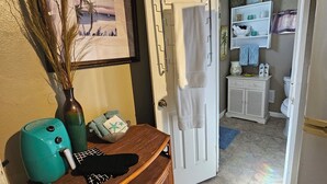 A hallway with a door leading to a bathroom, a framed picture on the wall, a basket with a towel, and a small table with a toaster and a vase.