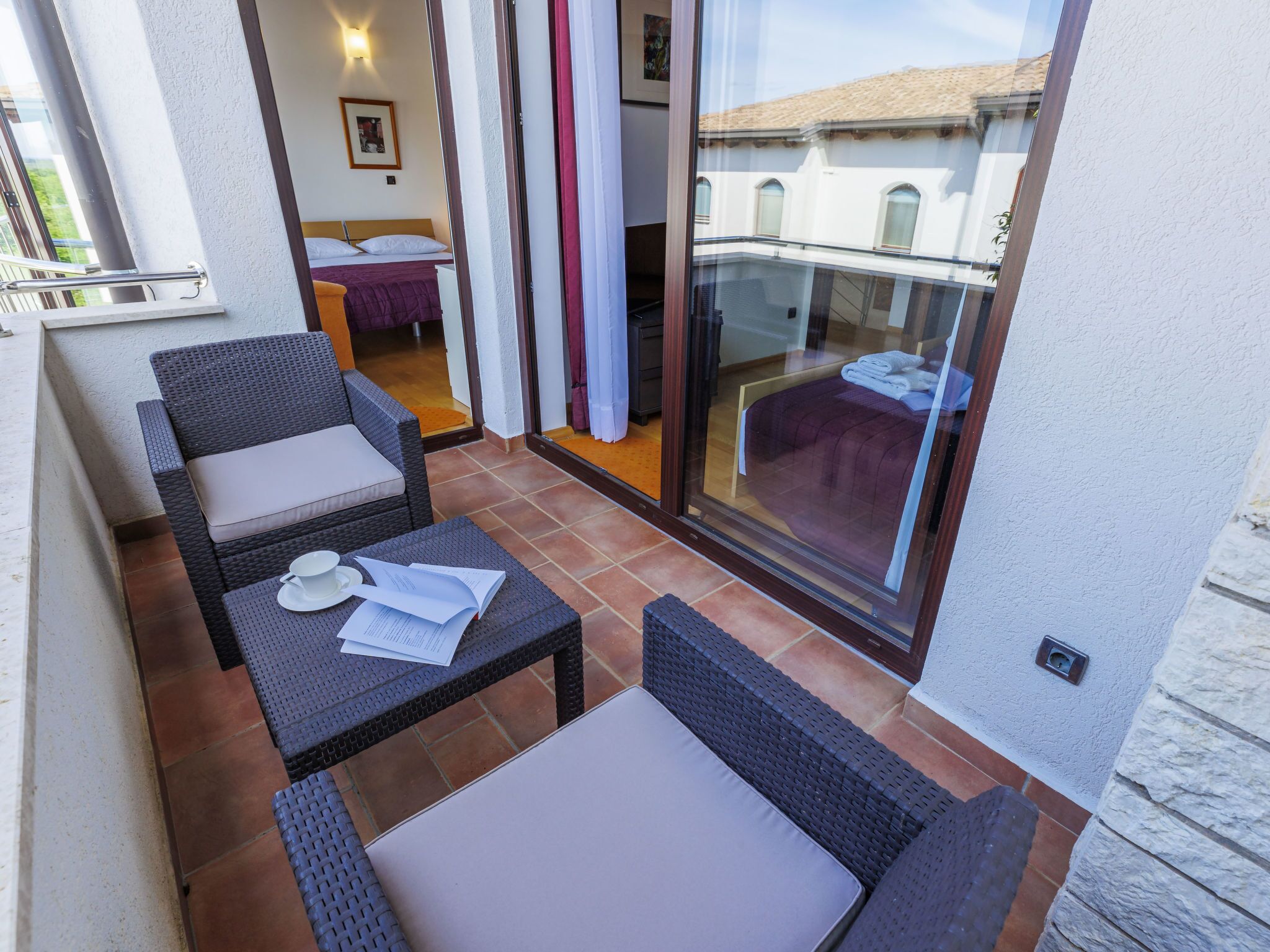 Un balcon avec des meubles en osier, une table recouverte de papiers et une vue sur un immeuble à travers une porte vitrée.