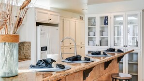 A kitchen with a marble countertop, wooden stools, white cabinets, and a vase with dried plants. - Massive Beach Home | Ocean View | Amazing Rooftop (San Diego)