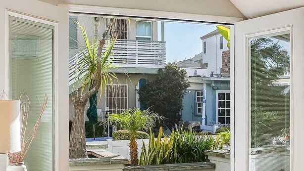 A modern living room with a brown sofa, a glass door leading to a balcony with plants and a view of neighboring houses. - Massive Beach Home | Ocean View | Amazing Rooftop (San Diego)