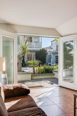 A modern living room with a brown sofa, a glass door leading to a balcony with plants and a view of neighboring houses.