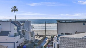 A view of a coastal residential area with modern buildings, a palm tree, and a beach in the background. - Massive Beach Home | Ocean View | Amazing Rooftop (San Diego)