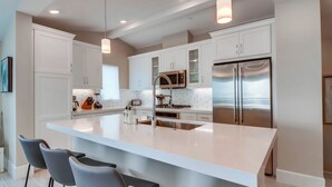 A modern kitchen with white cabinetry, a large island with bar stools, stainless steel appliances, and pendant lighting. - Steps to Water - Newly Built Bayside Dream | Patios, Fire Pit, Bay Views, Garage (San Diego)