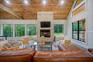 Ein geräumiges Wohnzimmer mit Kamin, Ledersofas und großen Fenstern mit Blick auf den Wald.