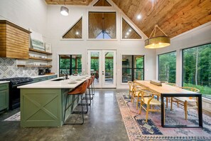 A modern kitchen with a large island, wooden ceiling, and large windows overlooking a forest.