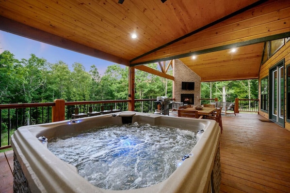 A hot tub on a wooden deck with a brick fireplace and outdoor furniture.