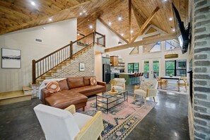 A spacious living area with a brown leather sofa, white armchairs, a coffee table, and a staircase leading to an upper level.