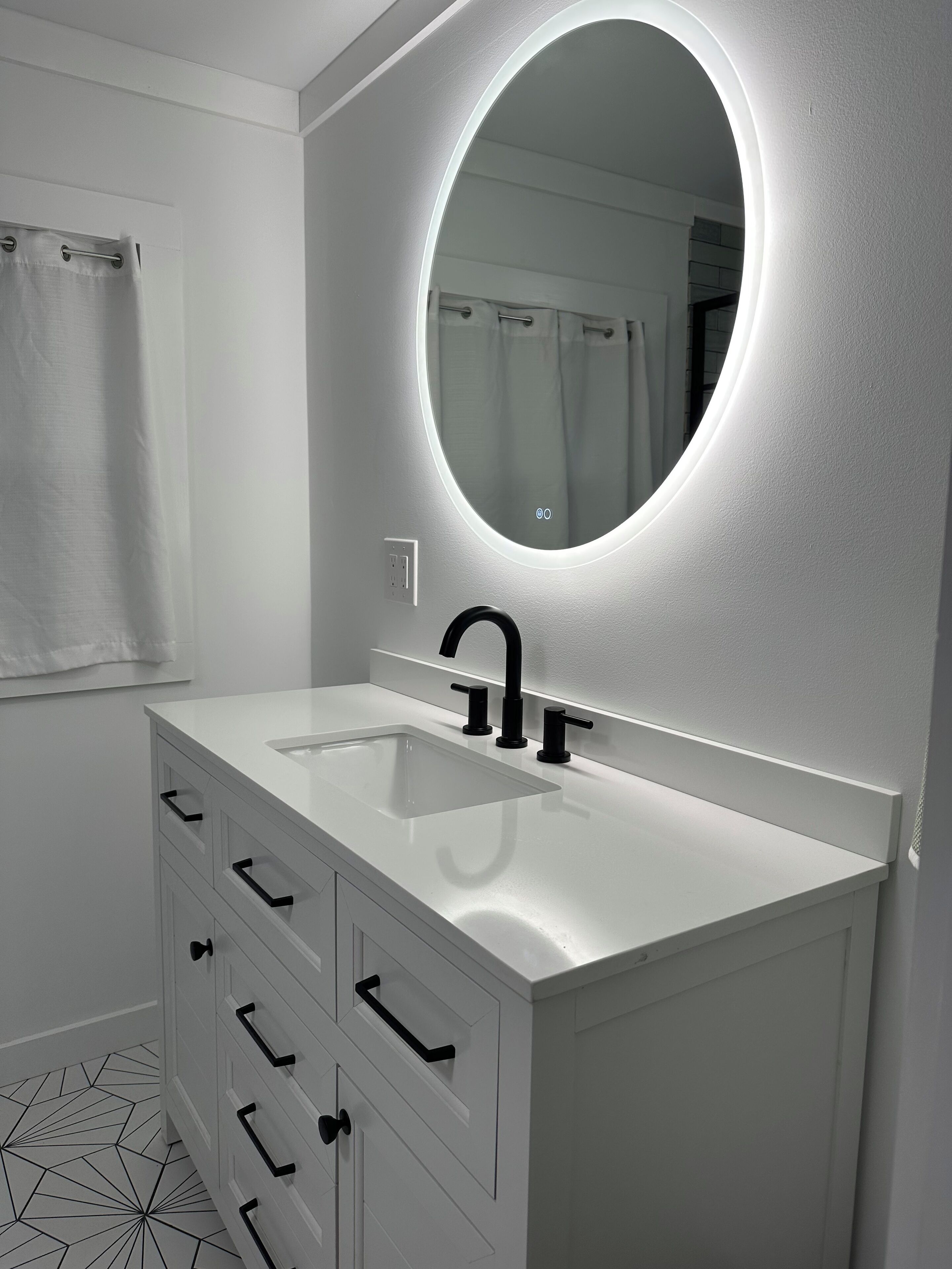 Un bagno moderno con un mobile lavabo bianco, uno specchio rotondo con illuminazione integrata, un rubinetto nero e un pavimento con motivo geometrico.