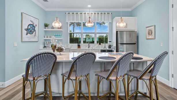 A modern kitchen with a central island, white cabinetry, and a stainless steel refrigerator. There are four wicker chairs around the island and a large window with white curtains. - Villa Miramar | Luxury 7-Bedroom Villa at Reunion Resort (Kissimmee)