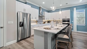 A modern kitchen with white cabinets, a stainless steel refrigerator, a central island with bar stools, and pendant lighting. - Villa Miramar | Luxury 7-Bedroom Villa at Reunion Resort (Kissimmee)