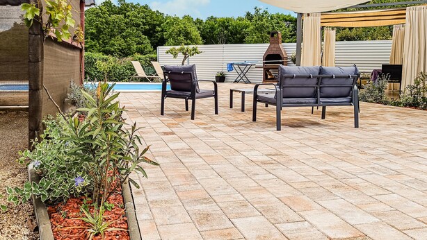 Eine Terrasse im Freien mit rotem Backsteinboden, zwei schwarzen Lounge-Stühlen, einem kleinen Tisch und einem Grill.