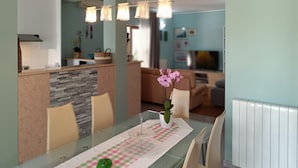 Ein Essbereich mit einem Glastisch, Stühlen und einer Vase mit Blumen. Im Hintergrund ist eine Küche mit einer Arbeitsplatte und einem Kühlschrank zu sehen.