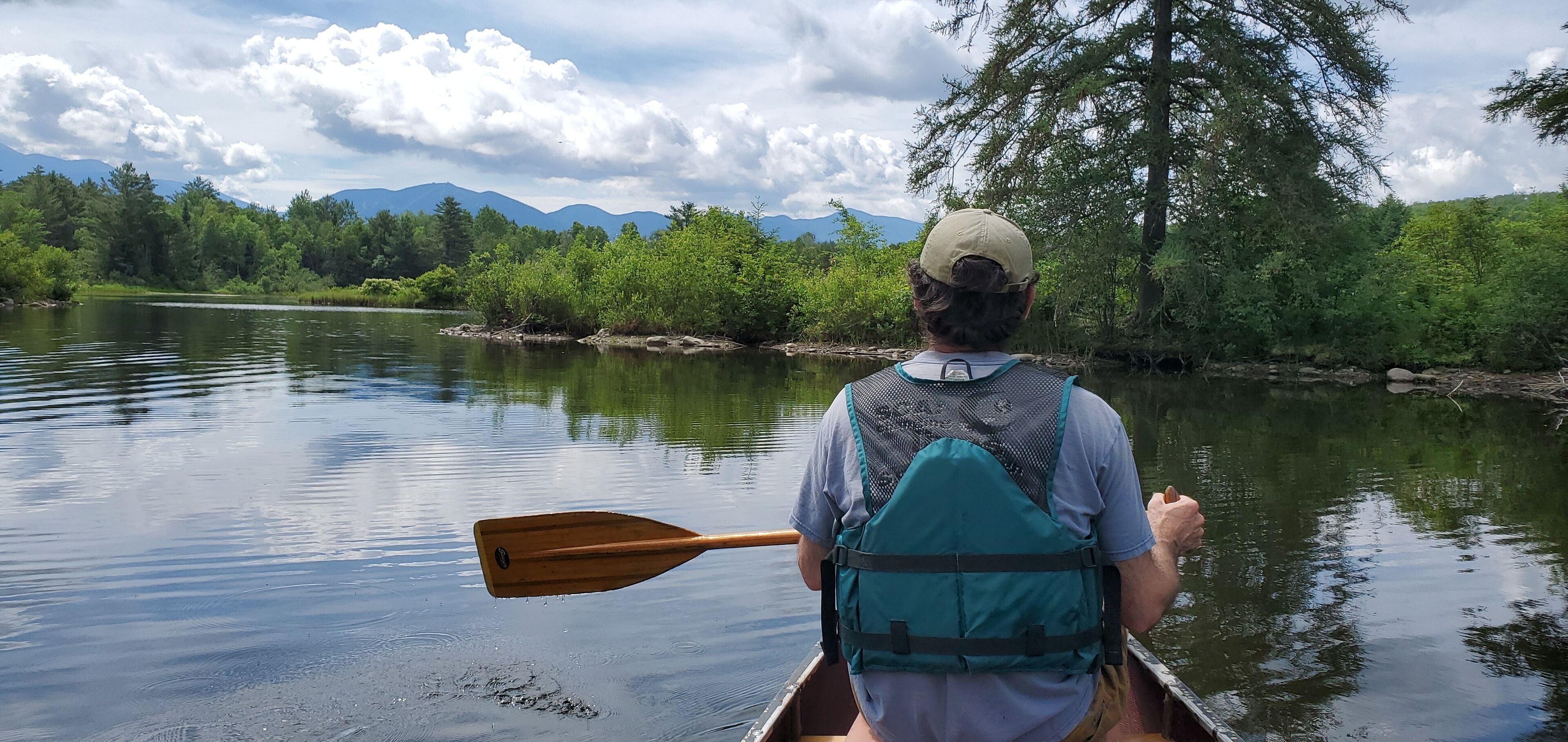 Una persona en una canoa sobre un río tranquilo, rodeada de vegetación exuberante y montañas al fondo.