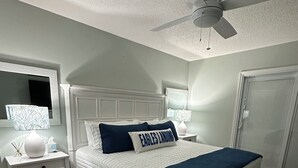 Een slaapkamer met een wit bed, een plafondventilator, een nachtkastje met een lamp en een deur.